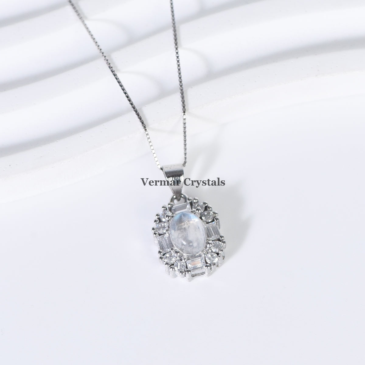Delicate silver pendant with oval blue moonstone surrounded by clear crystals on fine chain against white background
