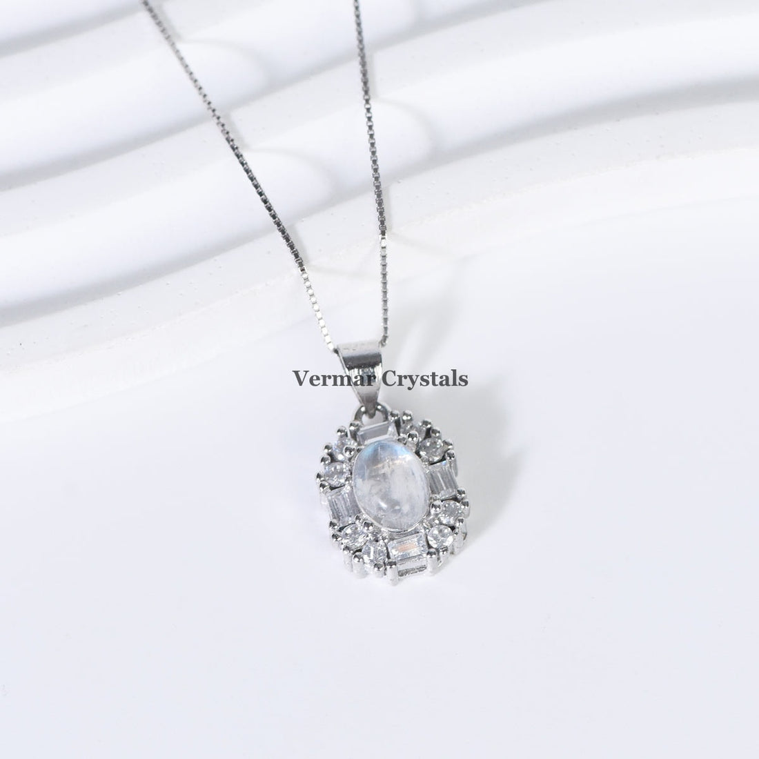 Delicate silver pendant with oval blue moonstone surrounded by clear crystals on fine chain against white background