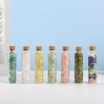 Seven small glass bottles filled with assorted natural crystal chips sealed with cork stoppers on white surface