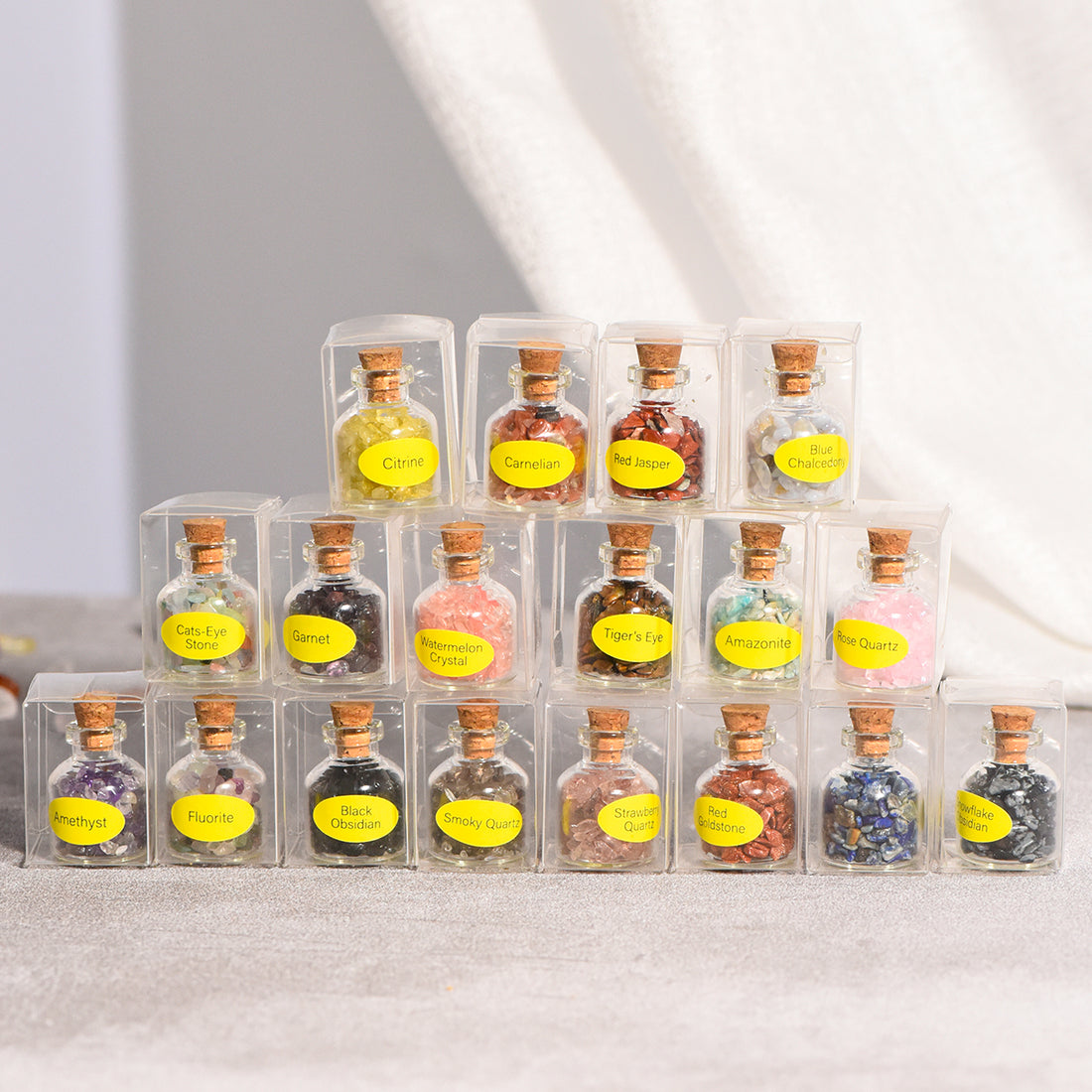 Stack of mini glass bottles with cork stoppers filled with assorted natural crystal chips in clear boxes on neutral background