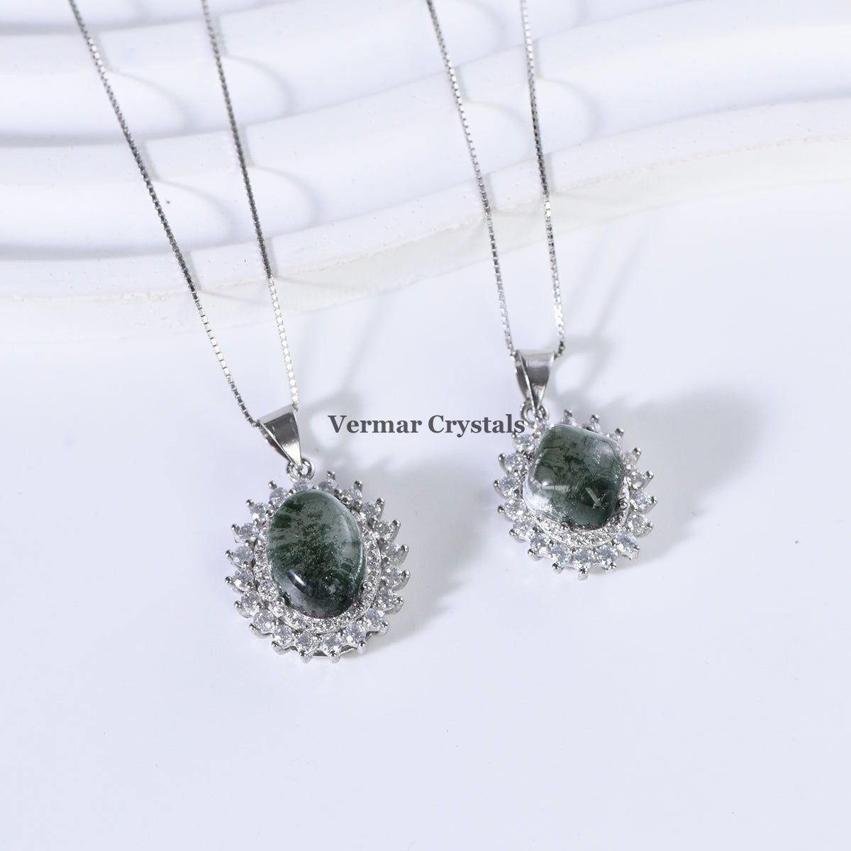 Two green phantom quartz pendants with silver halos and delicate chains on white textured surface