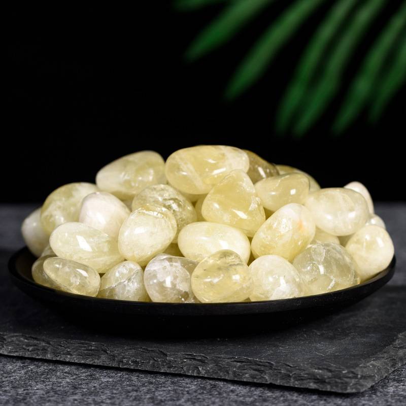 Polished yellow citrine tumbled stones piled on a black dish with dark textured background and green leaves above.