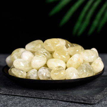 Polished yellow citrine tumbled stones piled on a black dish with dark textured background and green leaves above.