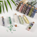 Eleven mini glass vials filled with colorful natural crystal chips with cork stoppers on white surface and green leaves background