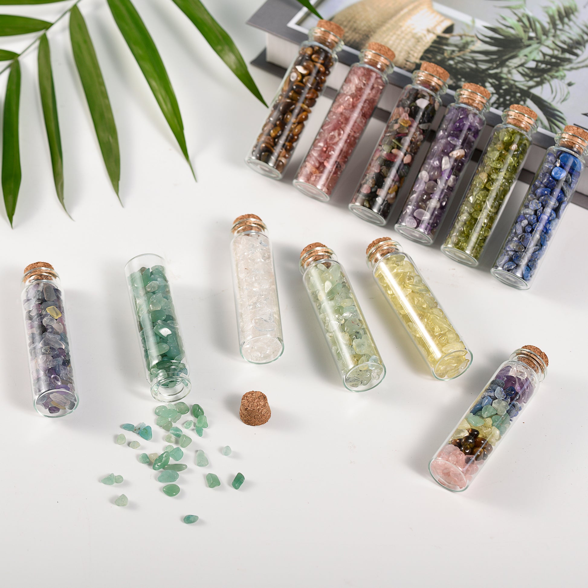 Eleven mini glass vials filled with colorful natural crystal chips with cork stoppers on white surface and green leaves background