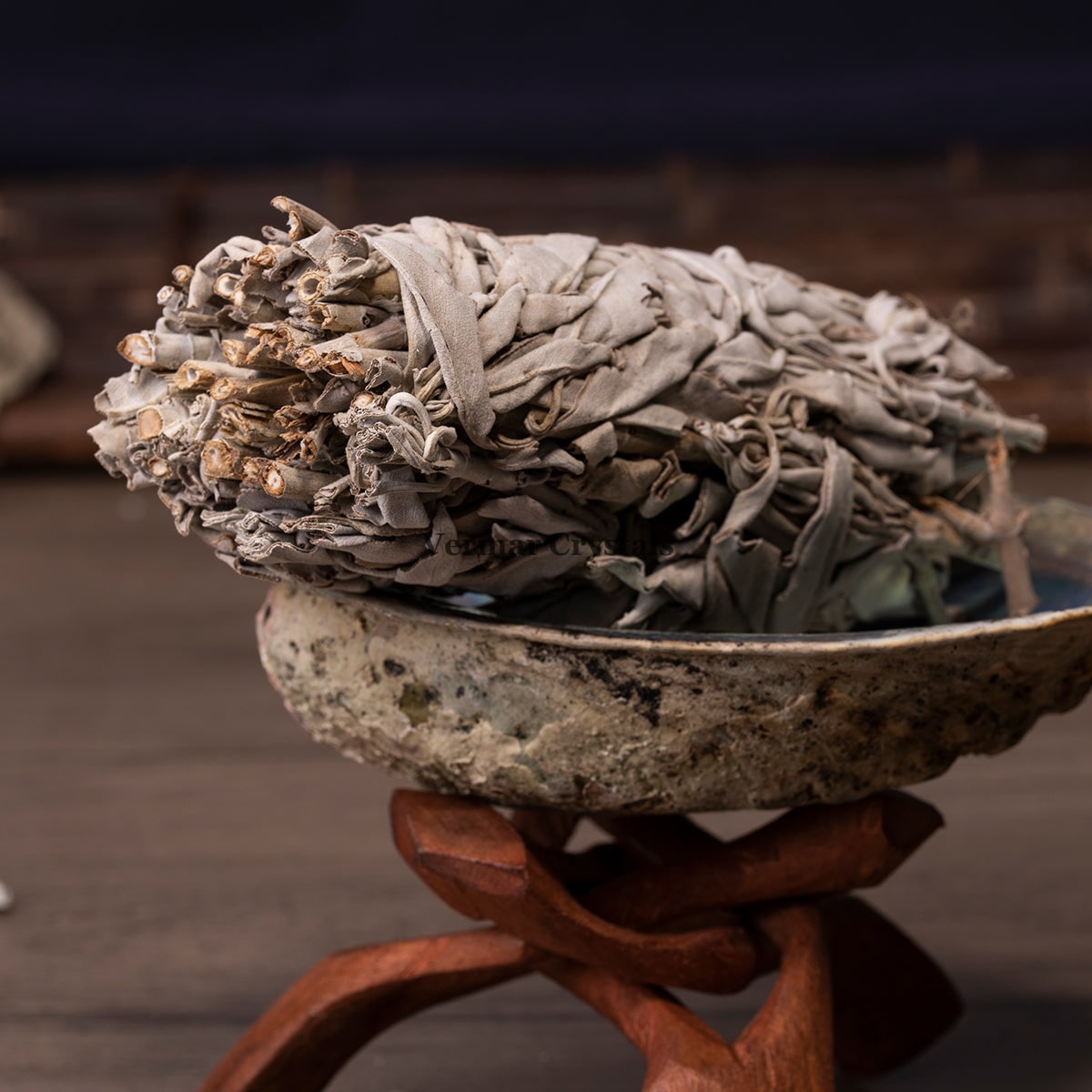 Close-up of dried white sage bundle resting in abalone shell on wooden stand for energy cleansing ritual