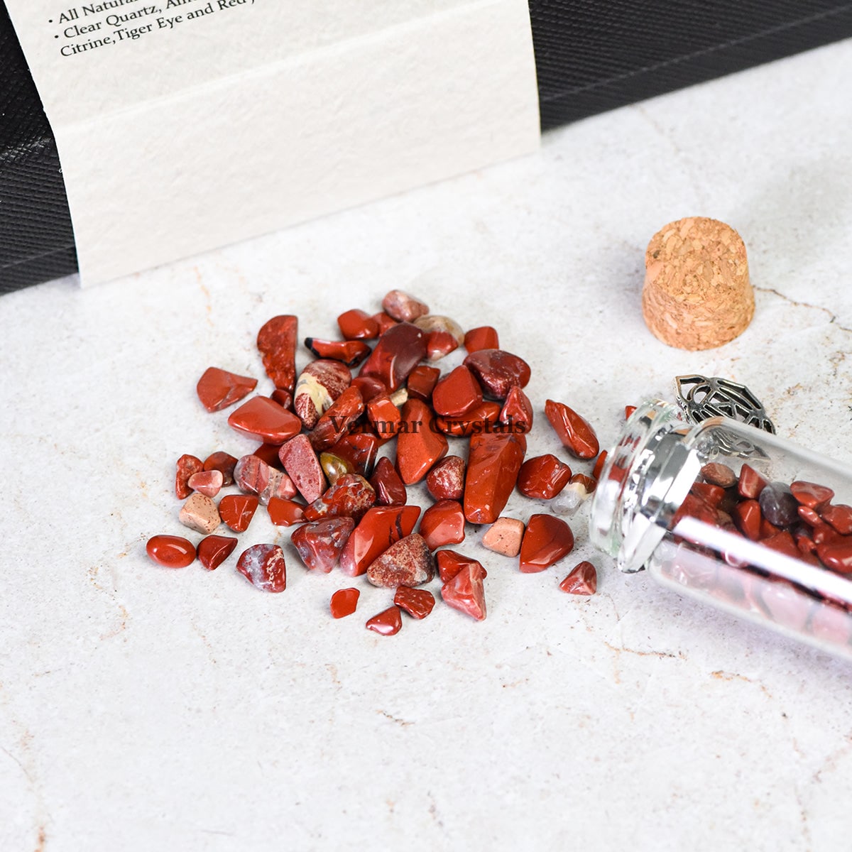 Close-up of red chakra healing chip stones spilling from a clear glass wishing bottle with a cork stopper on white surface
