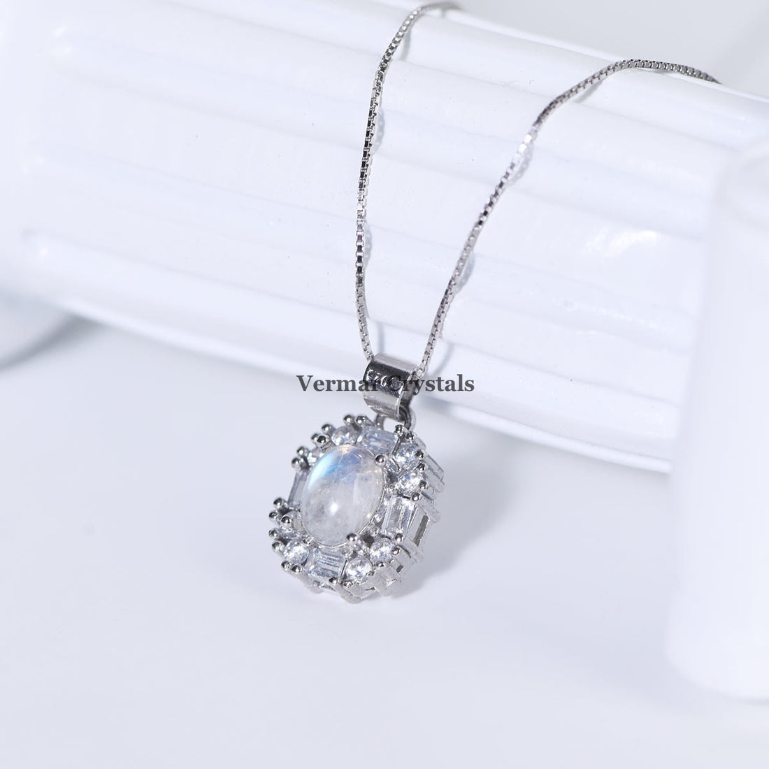 Delicate silver pendant with oval blue moonstone surrounded by clear crystals on fine chain against white background
