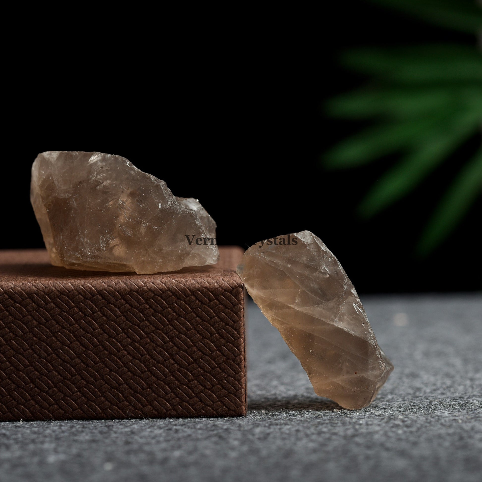 Two rough smoky quartz crystal stones displayed on textured brown and gray surfaces with dark background