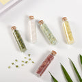 Five small glass vials filled with various colored natural crystal chips sealed with cork stoppers on white surface