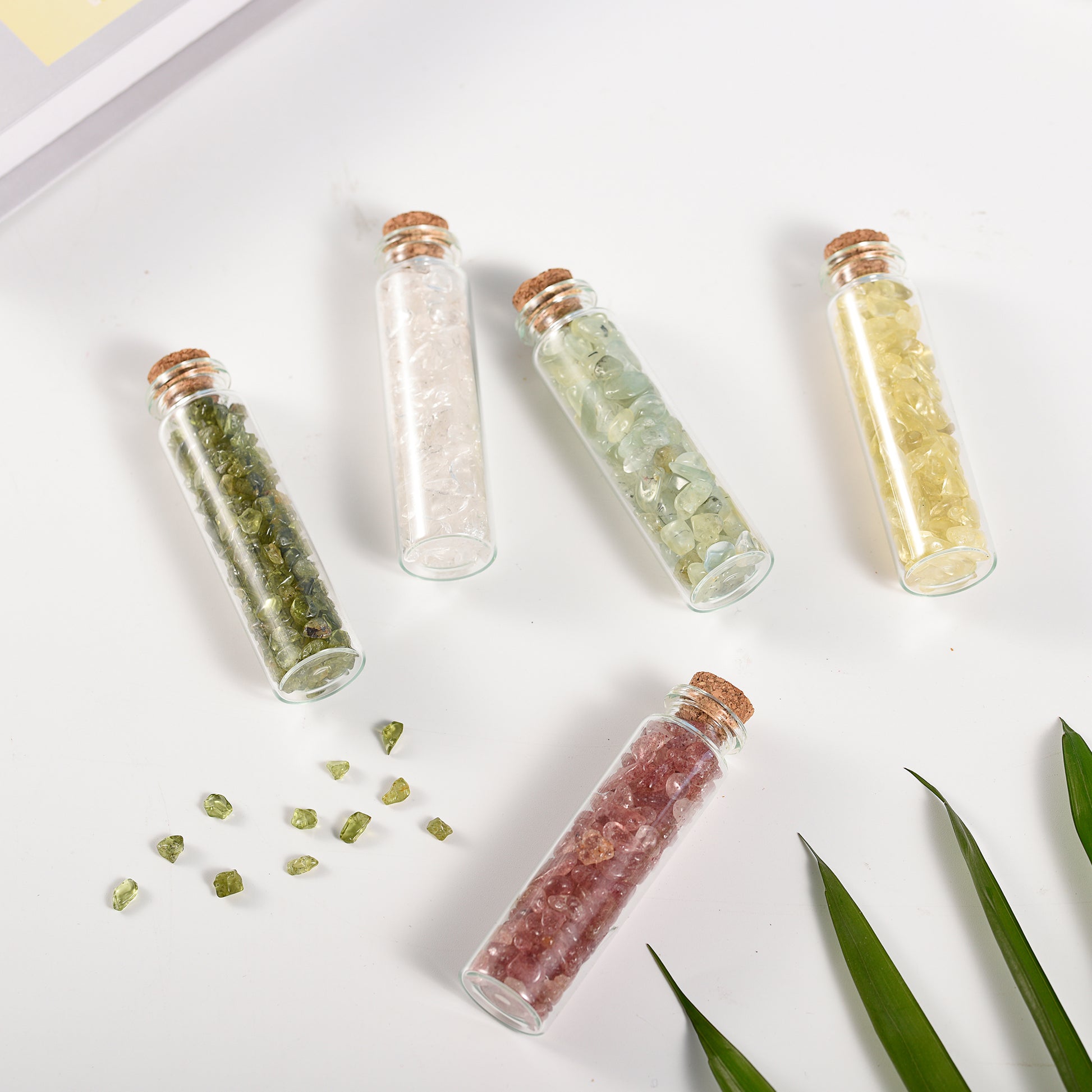 Five small glass vials filled with various colored natural crystal chips sealed with cork stoppers on white surface
