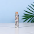 Clear glass vial filled with mixed natural crystal chips and sealed with a cork on clean surface with plant leaf background