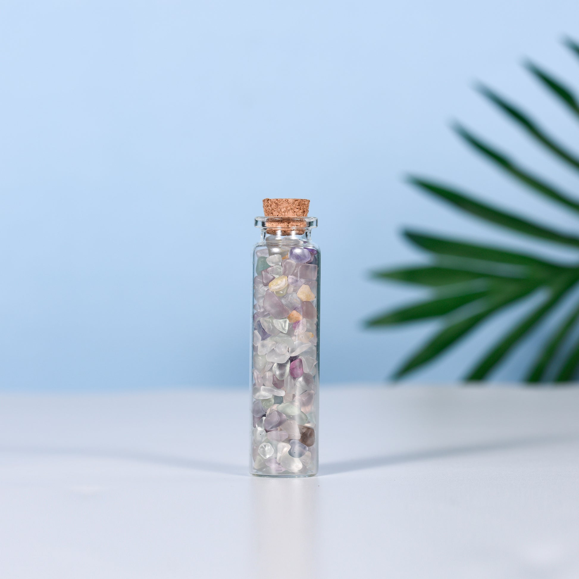 Clear glass vial filled with mixed natural crystal chips and sealed with a cork on clean surface with plant leaf background