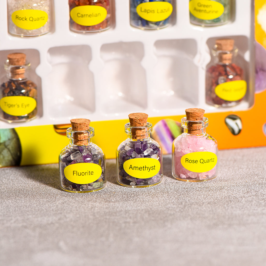 Stack of mini glass bottles with cork stoppers filled with assorted natural crystal chips in clear boxes on neutral background