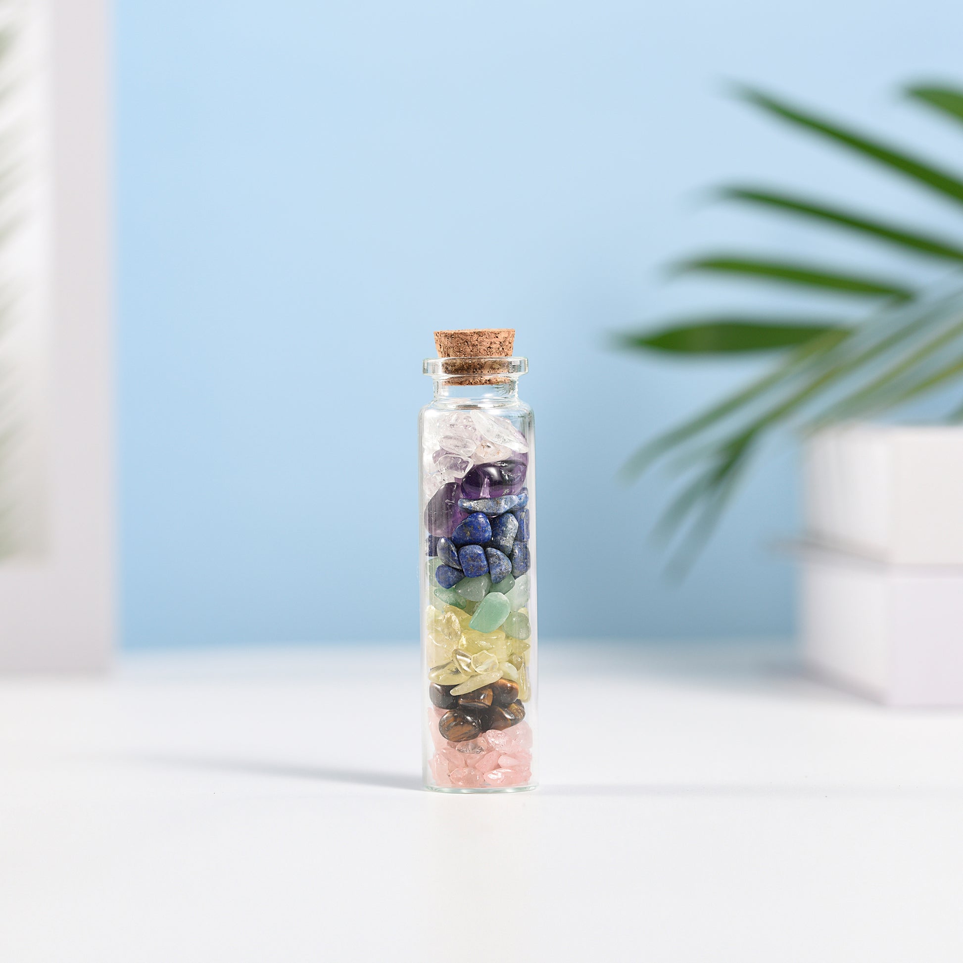 Mini glass vial filled with layered natural crystal chips and sealed with a cork on white surface with soft background