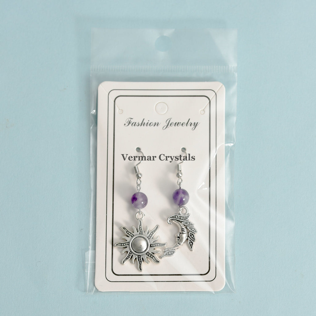 Asymmetric sun and moon silver earrings with polished amethyst beads hanging against white cup background