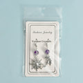 Asymmetric sun and moon earrings with amethyst beads in clear plastic packaging on light blue background