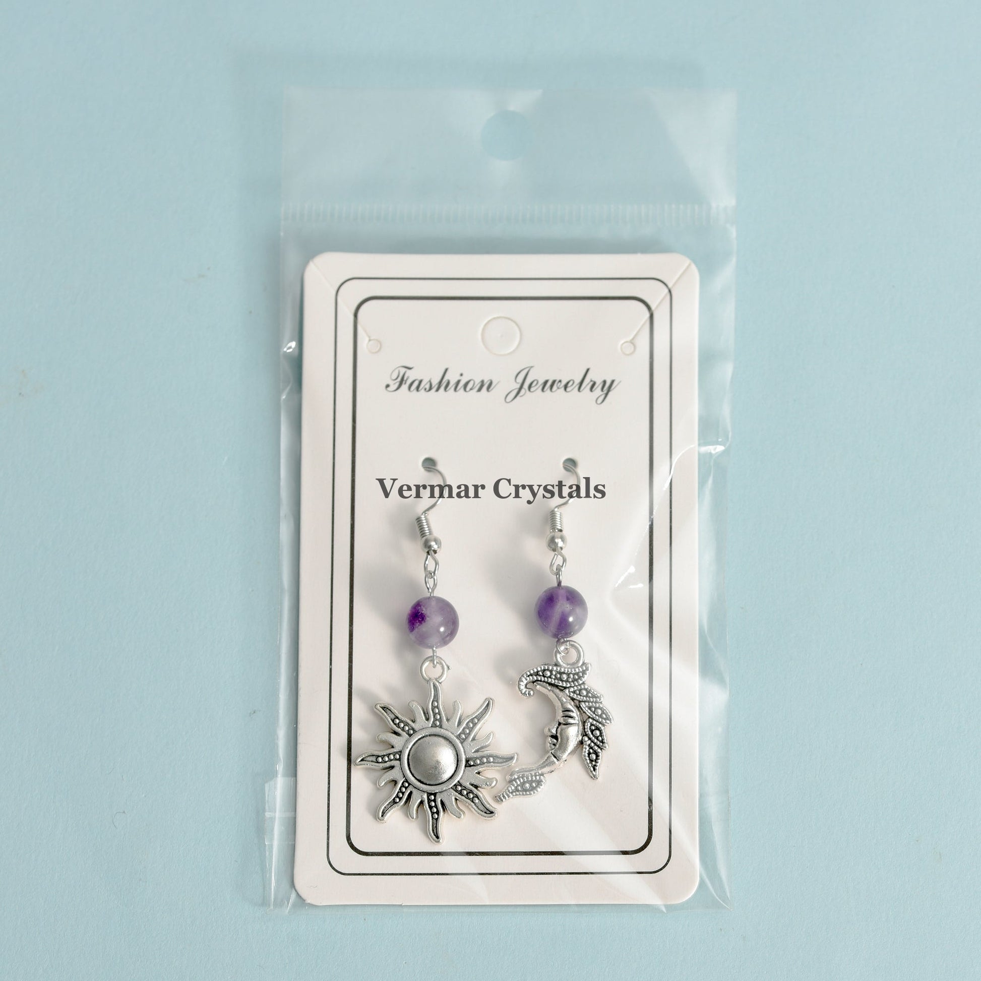 Asymmetric sun and moon earrings with amethyst beads in clear plastic packaging on light blue background
