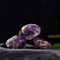 Three rough amethyst quartz crystal stones with purple and orange hues on dark fabric background