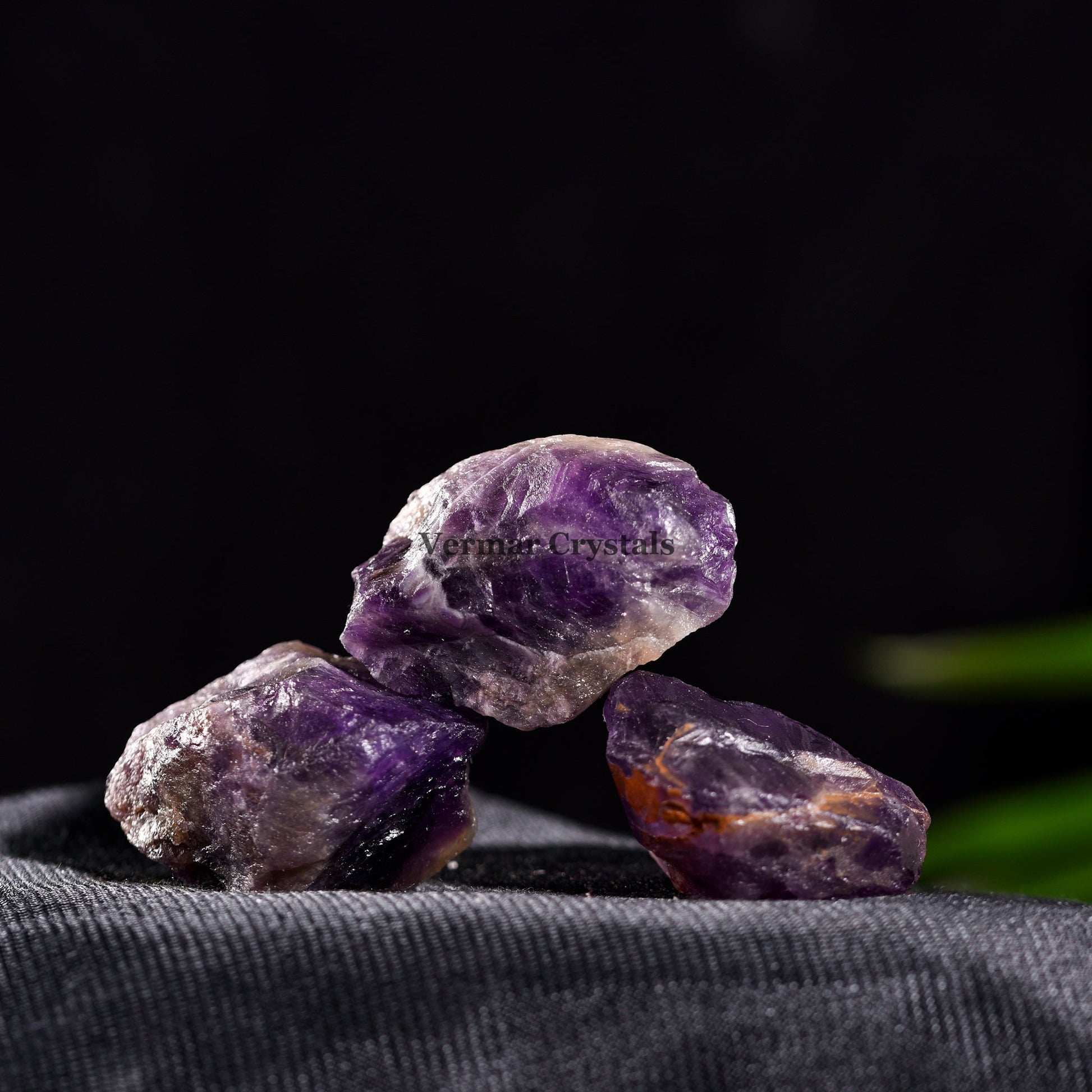 Three rough amethyst quartz crystal stones with purple and orange hues on dark fabric background
