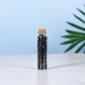Clear glass vial filled with small polished black natural crystal chips sealed with a cork stopper on white surface