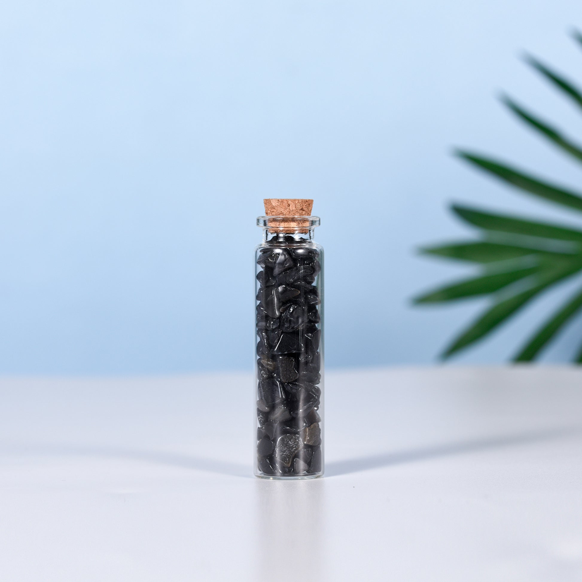 Clear glass vial filled with small polished black natural crystal chips sealed with a cork stopper on white surface