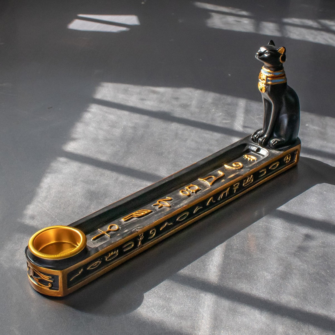 Black and gold Egyptian Guardian Cat incense holder with hieroglyphic details on gray surface with shadow patterns
