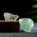 Two natural green fluorite crystal stones with translucent surfaces on a dark textured surface and box