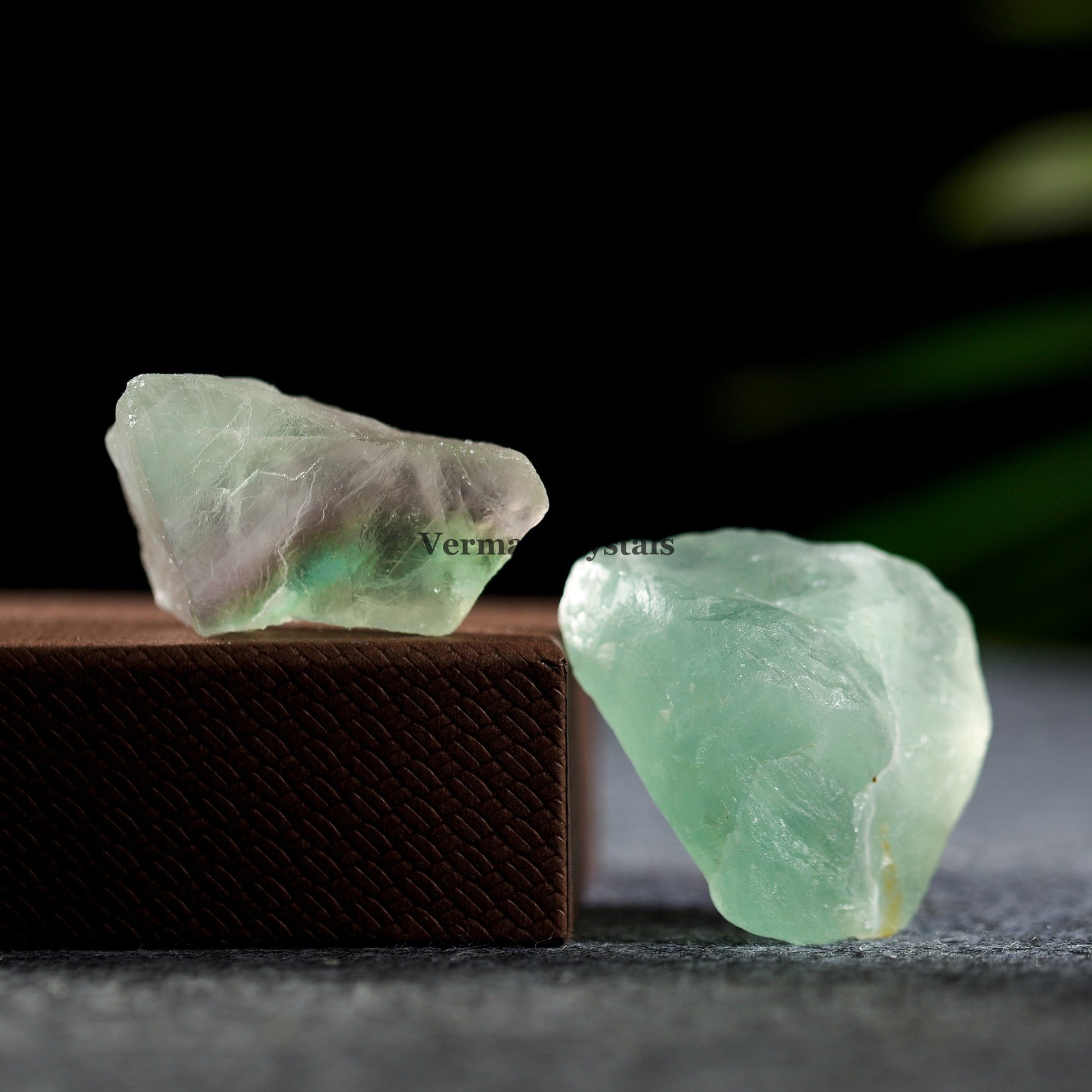 Two natural green fluorite crystal stones with translucent surfaces on a dark textured surface and box