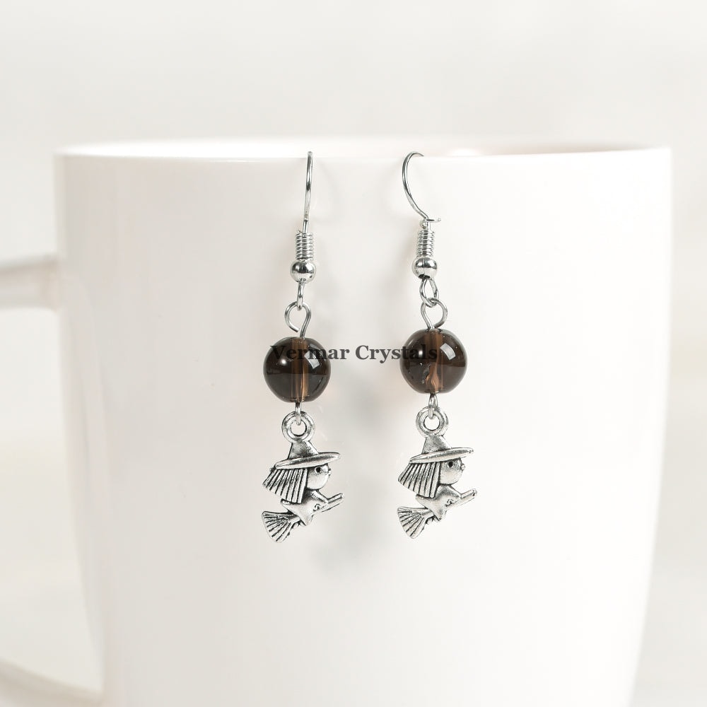 Pair of silver witch charm earrings with round smoky quartz beads hanging on a white ceramic cup background