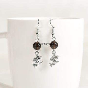 Pair of silver witch charm earrings with round smoky quartz beads hanging on a white ceramic cup background