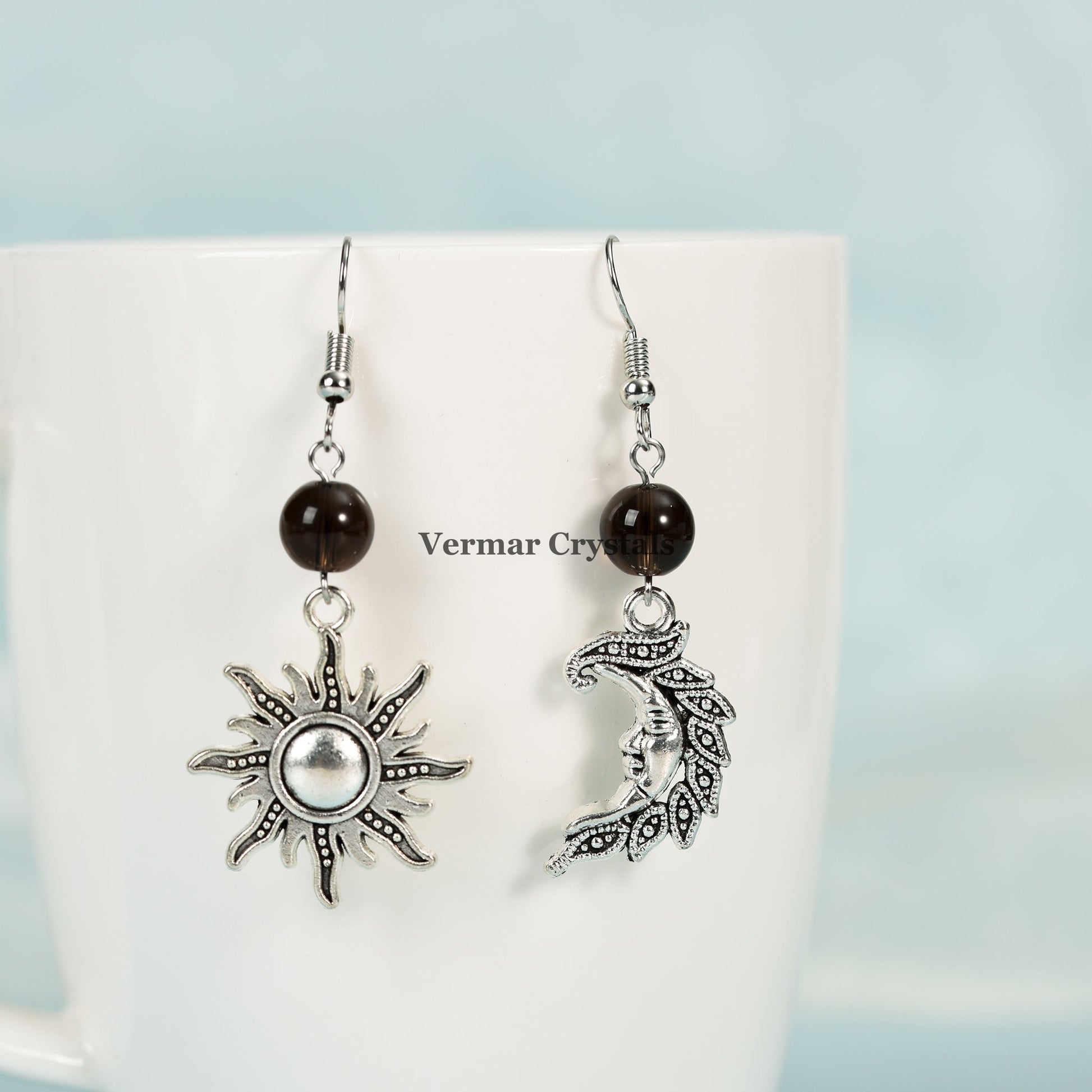 Asymmetric sun and moon earrings with smoky quartz beads and silver-tone metal charms hanging on white mug