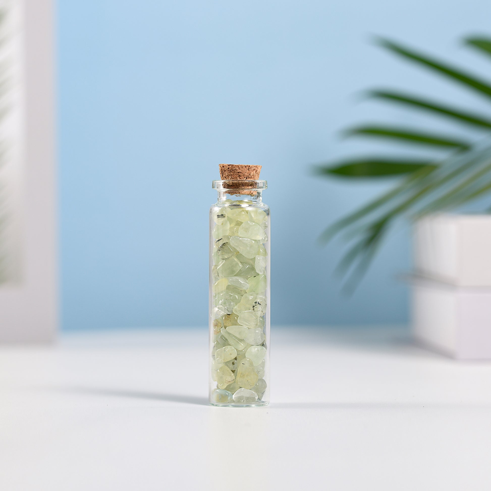 Mini glass vial filled with pale green natural crystal chips sealed with cork on white surface with soft background