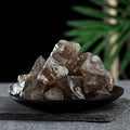 Pile of irregular translucent smoky quartz crystal stones on a black plate with dark background and green leaves