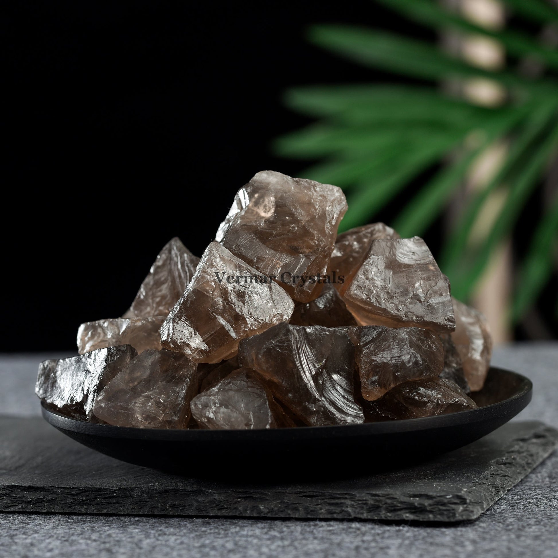 Pile of irregular translucent smoky quartz crystal stones on a black plate with dark background and green leaves