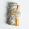 Bundle of dried sage, palo santo wood stick, and clear quartz crystal tied together with twine on white background