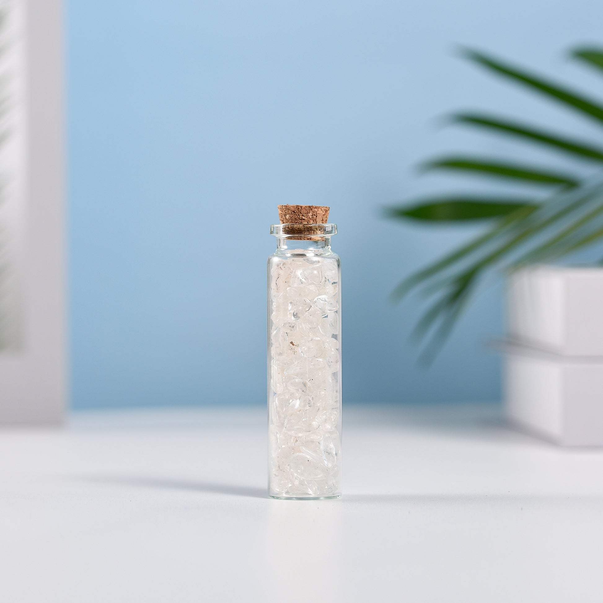 Mini glass vial filled with natural clear quartz crystal chips sealed with a cork on white surface and blue background