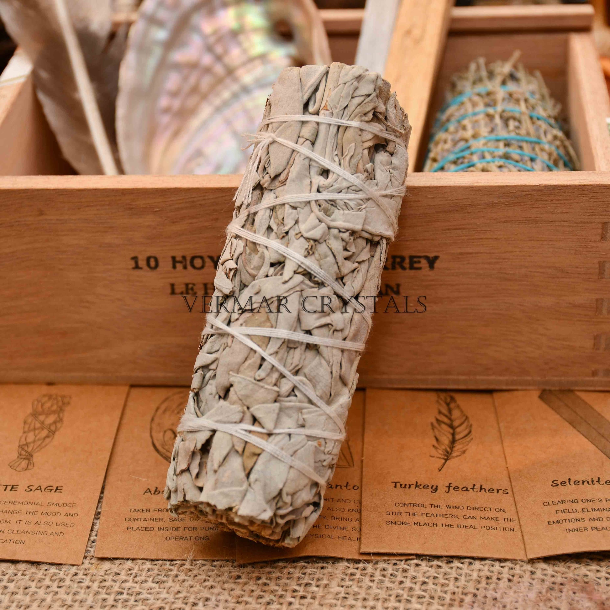 Bundle of dried white sage leaves wrapped with white string resting on wooden box with smudging items