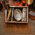 Wooden box containing abalone shell, selenite stick, palo santo, white sage bundle, and a brown feather on burlap surface
