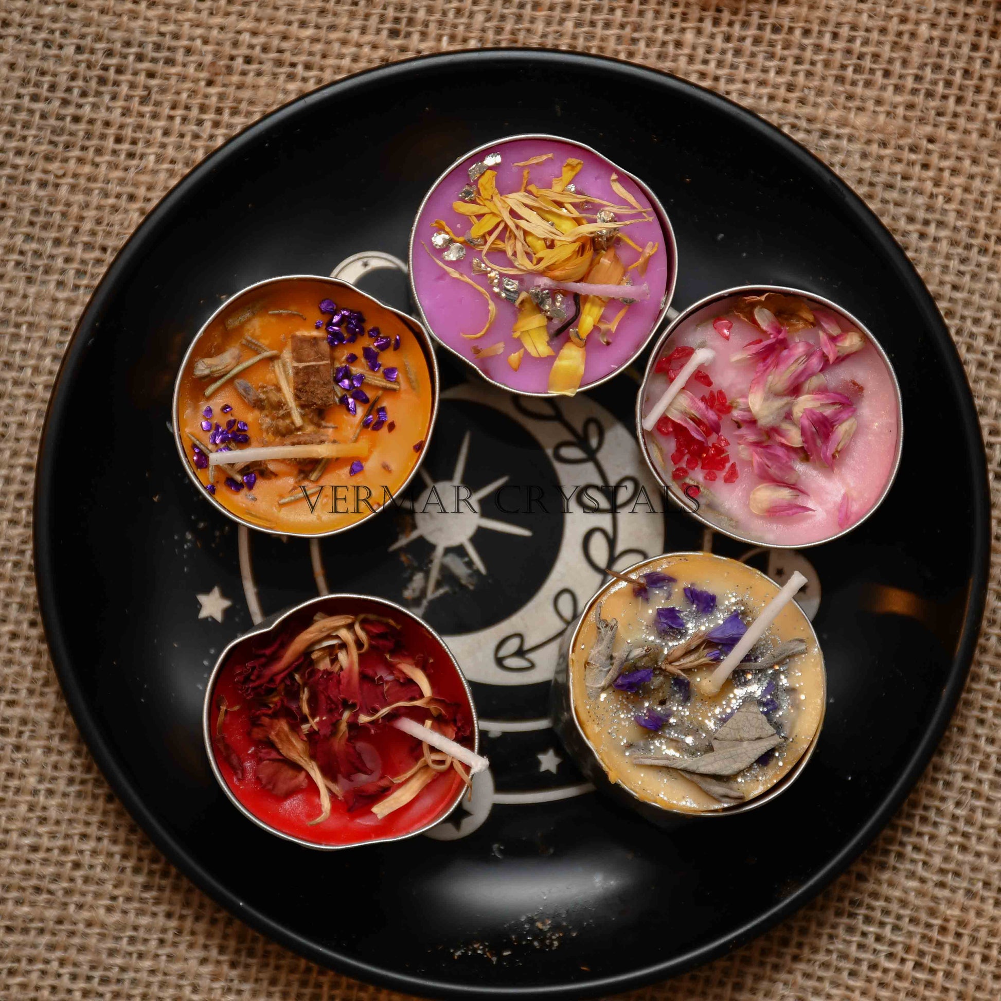 Five small colorful tealight candles with dried flower petals on black tray with moon and stars design on burlap background
