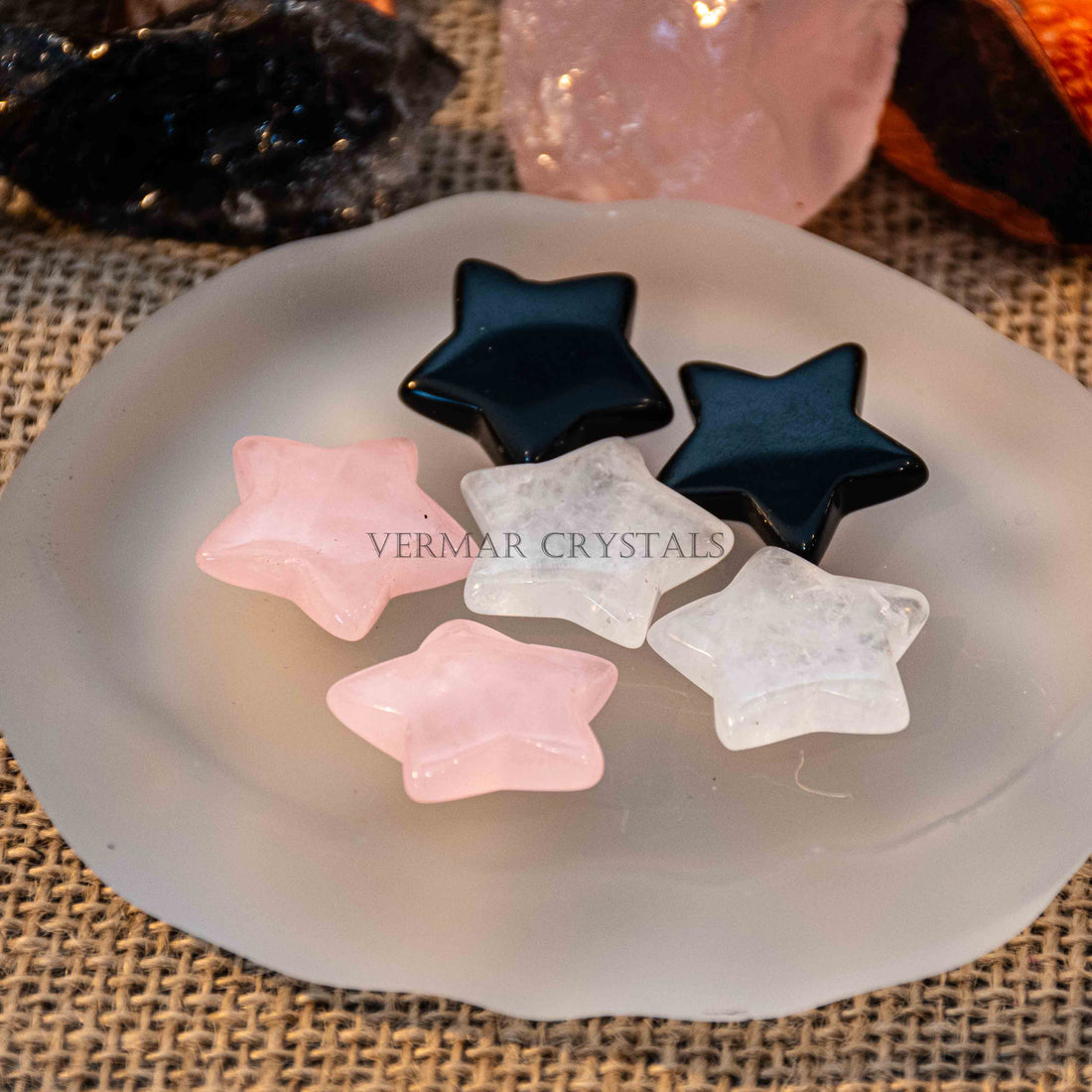 Mini pentagram-shaped tumbled stones in black, clear quartz, and rose quartz on frosted white dish with burlap background