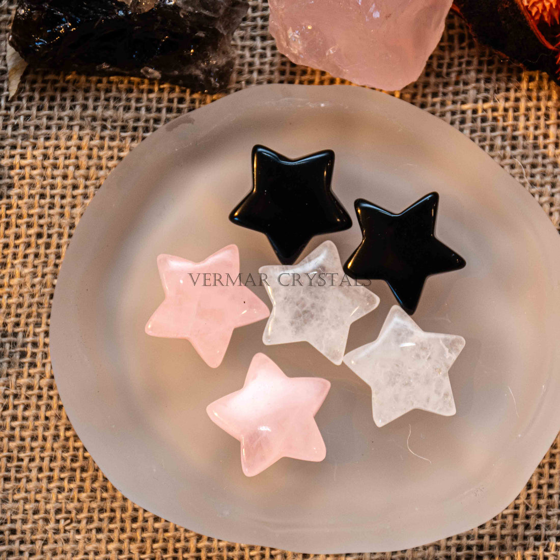 Mini pentagram-shaped tumbled stones in black, clear quartz, and rose quartz on frosted white dish with burlap background