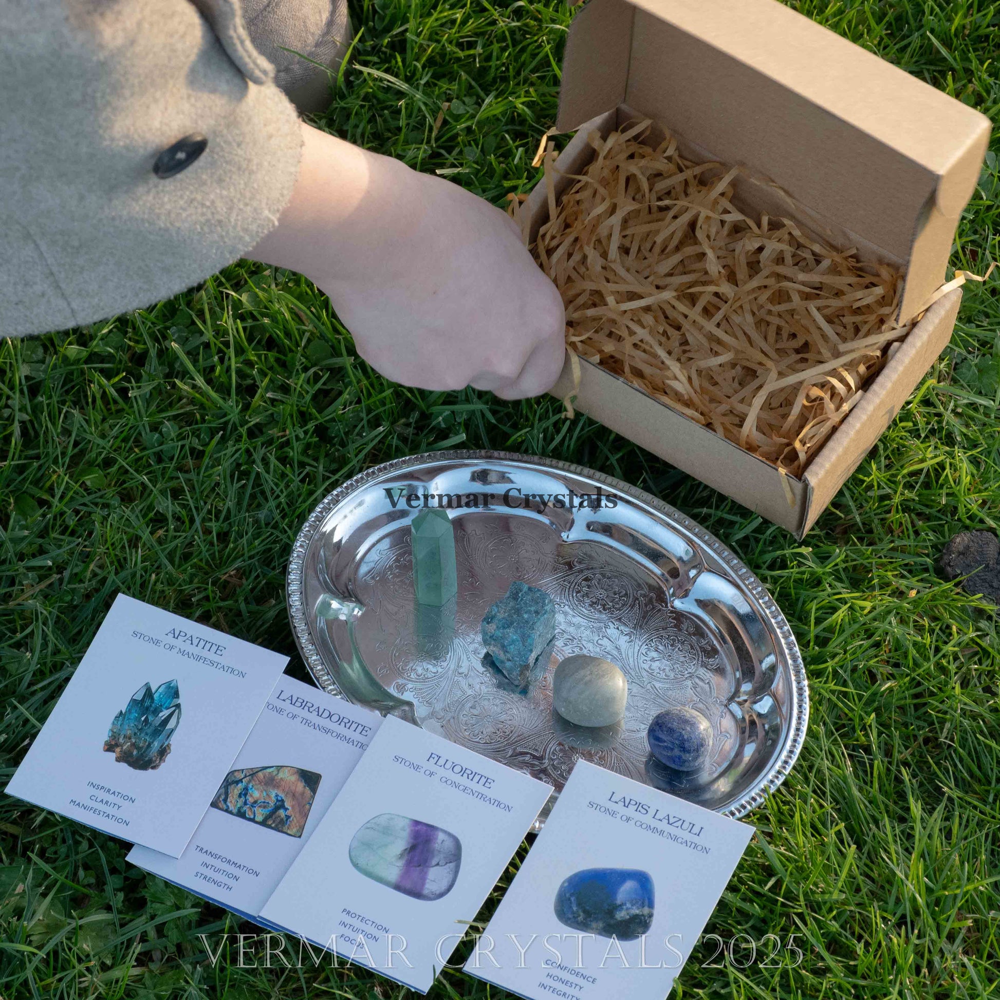 Hand placing natural crystals on silver plate alongside gemstone information cards and empty kraft box on grass background