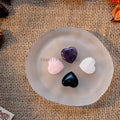 Four mini heart-shaped tumbled stones in amethyst, rose quartz, black onyx, and white selenite on a frosted dish