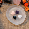 Four mini heart-shaped tumbled stones in various colors displayed on a smooth translucent dish on burlap background