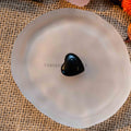 Mini heart-shaped black tumbled stone polished smooth, displayed on a round frosted white plate on burlap surface