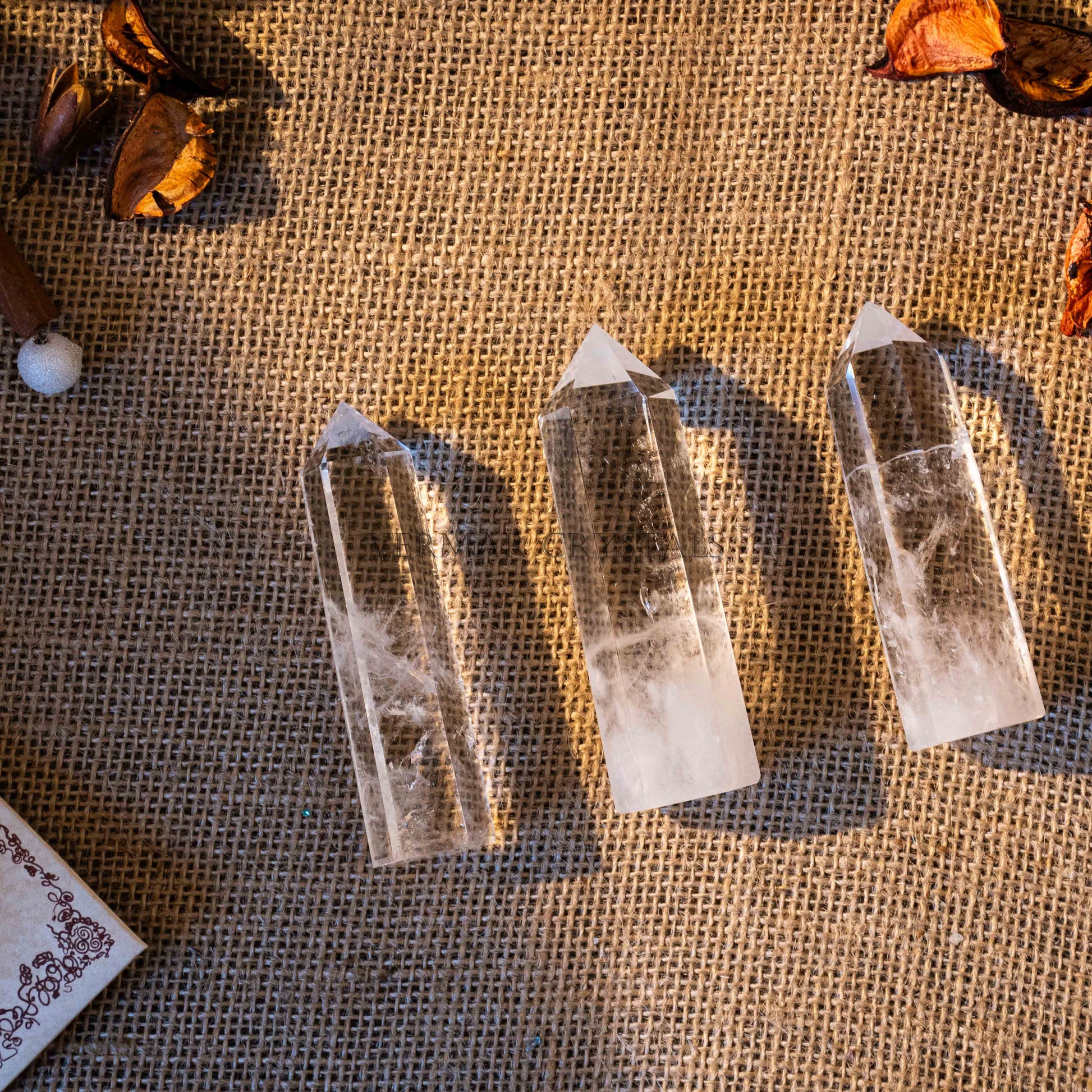 Three clear quartz crystal towers with pointed tips on textured burlap background with dried decorative elements
