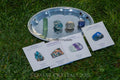 Set of four polished and raw crystals on silver tray with matching informational cards on green grass background