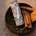 Premium white sage smudge stick with cinnamon sticks and dried herbs arranged in a round wooden bowl on burlap background
