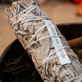 Close-up of a tightly wrapped white sage smudge stick with label on wooden bowl and blurred background